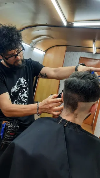 El barbero repasa con máquina un fade alto en la nuca, con el pelo largo peinado hacia atrás