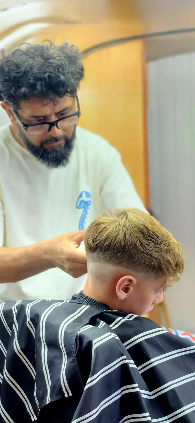 El barbero termina un fade medio con textura arriba en un cliente de pelo rubio