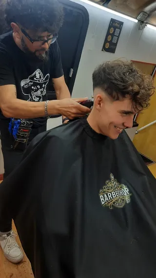 Un cliente sonríe mientras el barbero le marca con máquina un fade que deja los rulos largos arriba